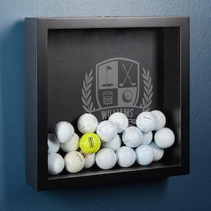 May include: Black shadow box with a golf-themed design and the name "WILLIAMS" engraved on it. The box is filled with golf balls, including one yellow golf ball.