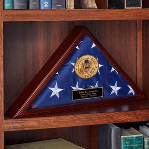 May include: A wooden triangular flag display case with a blue and white American flag inside. The flag has white stars and a gold Army emblem. A small plaque with the name "Brian F. Thompson" is visible.