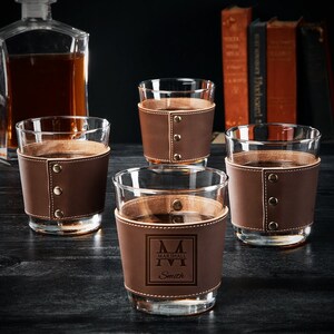 May include: Four brown leather wrapped glasses with a monogrammed initial "M" and the name "Marshall Smith" on one glass. The glasses are on a dark wood surface.