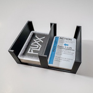 May include: A black card holder with two compartments, one holding a deck of cards labeled "FLUXX" and the other with cards featuring "PASS 1 LEFT" text. The holder is designed to organize and display card games.