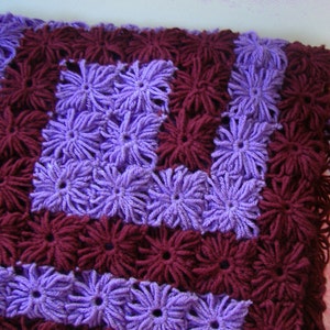 May include: A crocheted blanket with a purple and burgundy pattern. The blanket is made of yarn and has a textured surface.