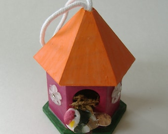 May include: A small, pink birdhouse with an orange roof and a white string for hanging. The birdhouse has a small opening for a bird to enter and a green base. A small bird is perched on the edge of the opening.