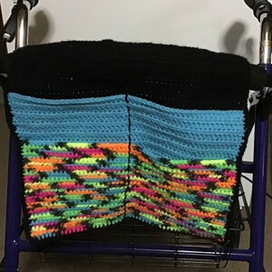 May include: A black, blue, and multi-colored crocheted bag is attached to a walker. The bag has two compartments.