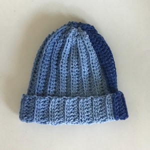 May include: A handmade, ribbed crochet beanie in shades of blue. The hat features a folded brim and a vertical ribbing pattern. The color transitions from light blue to a darker blue at the top and on the brim.