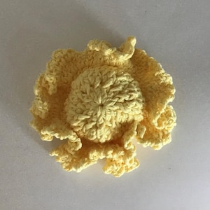 Könnte beinhalten: Handgefertigte, hellgelbe Häkelblume mit strukturiertem, gerüschtem Design. Die Blume hat ein zentrales, kreisförmiges Element, das von geschichteten Blütenblättern umgeben ist, wodurch ein dreidimensionaler Effekt entsteht. Die Häkelarbeit ist sichtbar.
