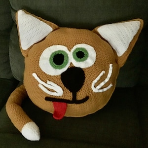 Puede incluir: Una almohada de gato de crochet con un cuerpo marrón, orejas blancas, ojos verdes, una nariz negra y una lengua roja que sobresale.