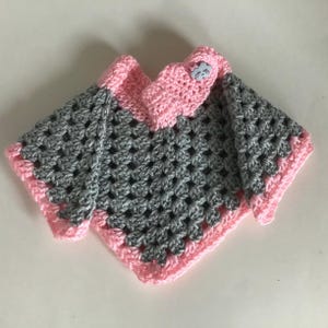 May include: A gray and pink crocheted baby sweater with a small cloud-shaped button. The sweater has a V-neck and a pink trim.