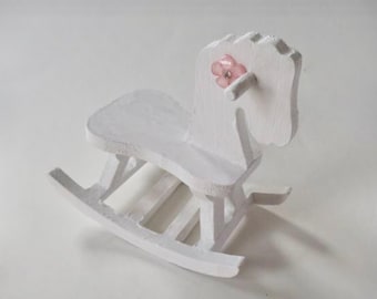 May include: White wooden rocking horse with a pink flower on its head. The rocking horse is a children's toy.
