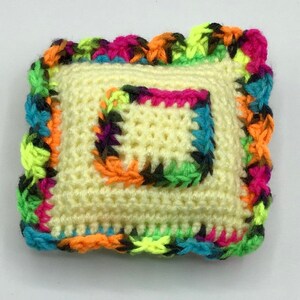 May include: A small, yellow crocheted pillow with a colorful crocheted border. The border is made of multiple colors, including pink, blue, green, and black.