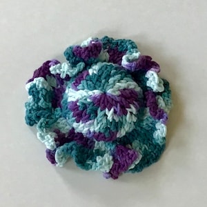 May include: A crocheted flower in shades of teal, purple, and white. The flower has a ruffled edge and is made with a variety of stitches.