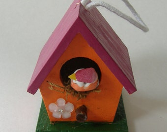 May include: A small wooden birdhouse with a pink roof, orange body, and a green base. A small bird with pink and yellow wings is inside the house.