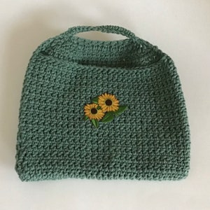 Könnte beinhalten: Eine grüne gehäkelte Einkaufstasche mit einem Sonnenblumenmuster.