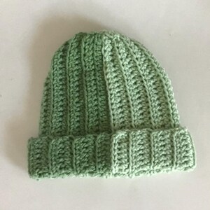 May include: A light green, ribbed crochet beanie hat. The hat is made of soft yarn and features a folded brim. The crochet stitches create a textured, vertical pattern across the hat's surface. The hat is a solid color and appears handmade.