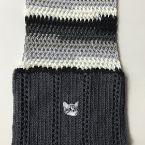 May include: A gray and white striped crocheted scarf with a black crocheted section at the bottom. A small gray and white cat face is embroidered on the black section.