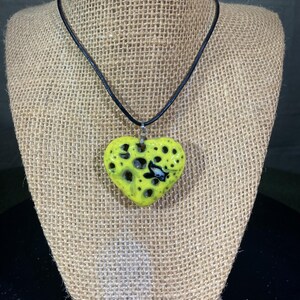 May include: A handmade necklace featuring a bright yellow heart-shaped pendant with black spots and a small black and white design. The pendant hangs from a black cord. The necklace is displayed on a burlap surface.