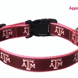 May include: A maroon and pink dog collar with the letters "A M" repeated in white. The collar has a black plastic buckle and a silver metal ring. The text "Aggies Pink" is visible in the upper right corner of the image.