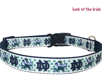 May include: A dog collar with a white ribbon featuring a repeating pattern of a leprechaun, the letters "ND", and green shamrocks. The collar has a navy blue backing and a black plastic buckle.