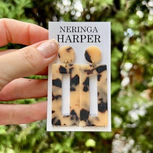 May include: A pair of dangle earrings featuring a circular stud and a rectangular drop piece, both displaying a beige and black tortoiseshell pattern. The earrings are attached to a white display card printed with the text NERINGA HARPER.