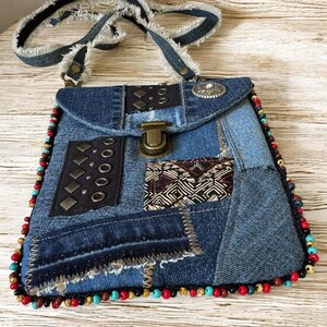Op de afbeelding: Een denim patchwork crossbody tas met een klepsluiting en een gefranjerde denim riem. De tas heeft decoratieve metalen studs, een patroonstof accent en een kralenrand in rood, turquoise en goud.