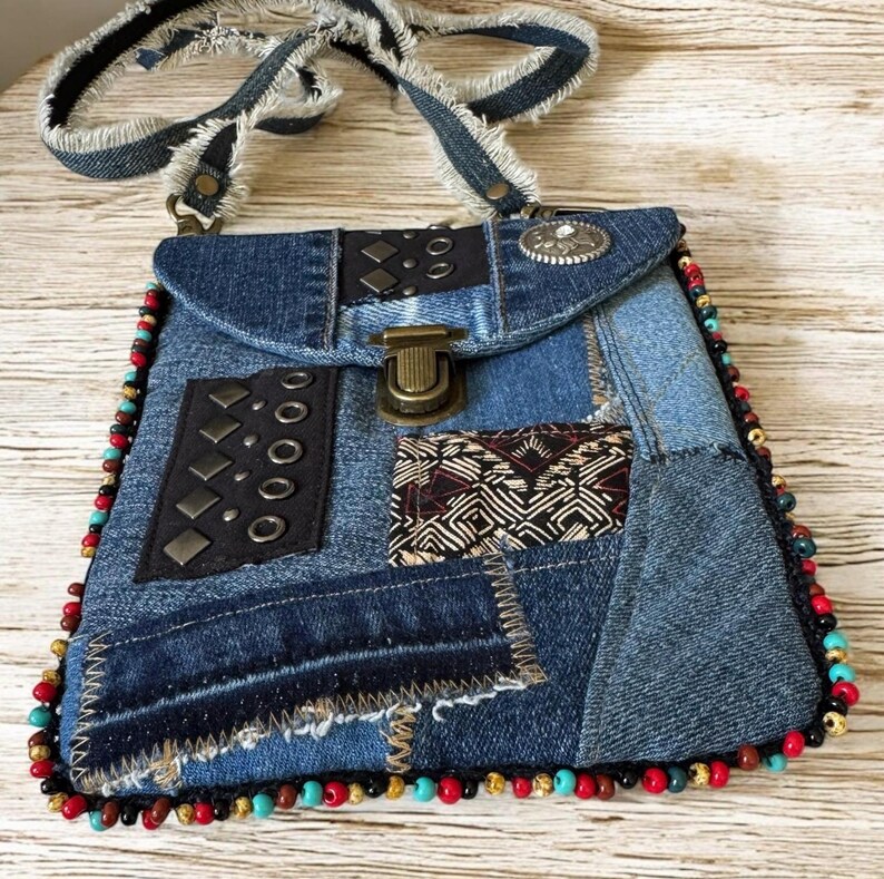 Op de afbeelding: Een denim patchwork crossbody tas met een klepsluiting en een gefranjerde denim riem. De tas heeft decoratieve metalen studs, een patroonstof accent en een kralenrand in rood, turquoise en goud.