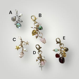 May include: Five decorative keychains with various charms, including seashells, pearls, crystals, and crosses. Each keychain has a lobster clasp and a collection of beads and pendants in different shapes and colors. The keychains are displayed against a neutral background.