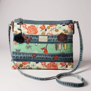 May include: A patchwork crossbody bag with a denim strap. The bag features floral and cat print fabric panels, accented with denim strips. A butterfly charm and a black pom-pom hang from the zipper. The bag has a rectangular shape.