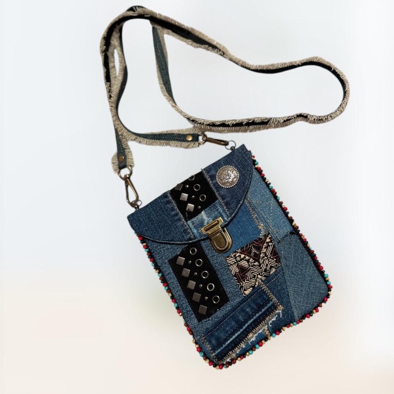 Op de afbeelding: Een denim crossbody tas met een patchwork design. De tas heeft een verscheidenheid aan denim tinten, versierd met zwarte studs en een decoratieve metalen sluiting. De riem is gemaakt van denim en een beige stof. De randen zijn afgewerkt met kleurrijke kralen.