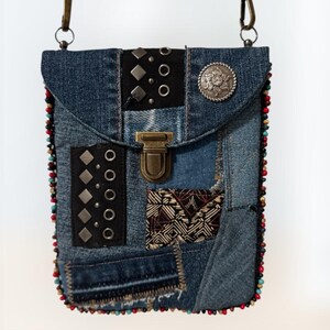 Op de afbeelding: Een denim crossbody tas met een patchwork design. De tas heeft zwarte leren accenten met zilveren studs en een decoratieve metalen knoop. De randen zijn afgewerkt met kleurrijke kralen en hij heeft een metalen sluiting en een schouderriem.