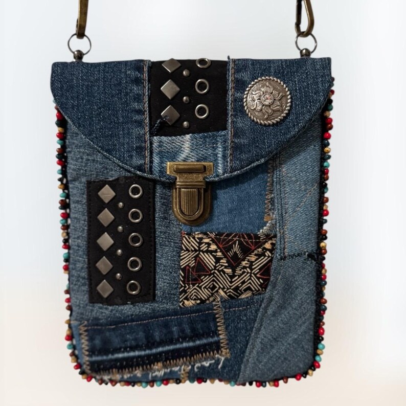 Op de afbeelding: Een denim crossbody tas met een patchwork design. De tas heeft zwarte leren accenten met zilveren studs en een decoratieve metalen knoop. De randen zijn afgewerkt met kleurrijke kralen en hij heeft een metalen sluiting en een schouderriem.