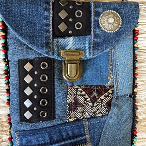 Op de afbeelding: Een denim patchwork crossbody tas met een klepsluiting. De tas heeft zwarte leren accenten met zilveren studs en een decoratieve zilveren knoop. De randen zijn afgewerkt met kleurrijke kralen en de tas heeft een messing sluiting.