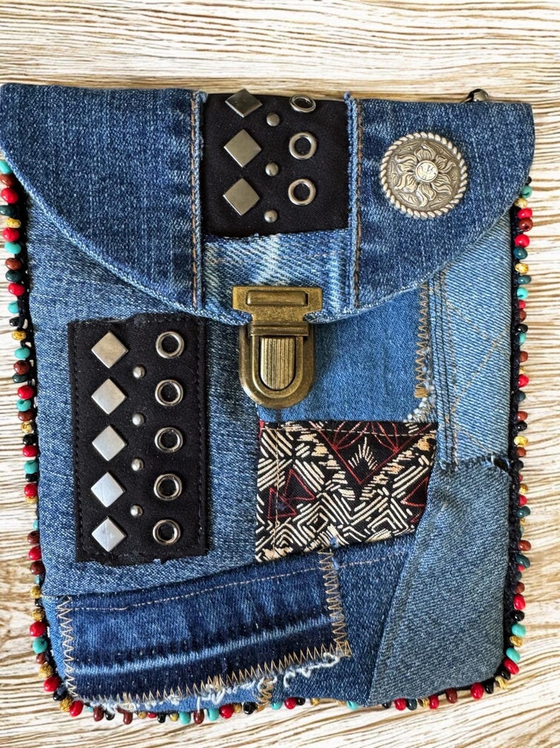 Op de afbeelding: Een denim patchwork crossbody tas met een klepsluiting. De tas heeft zwarte leren accenten met zilveren studs en een decoratieve zilveren knoop. De randen zijn afgewerkt met kleurrijke kralen en de tas heeft een messing sluiting.