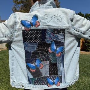 Puede incluir: Chaqueta vaquera azul claro con mariposas azules bordadas y detalles de patchwork en la espalda. Las mariposas tienen alas azules con acentos rojos y rosas. El patchwork presenta varios tonos de azul.