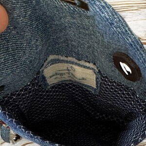 Op de afbeelding: Een denim handtas met een donkerblauwe binnenkant en een wit gestikt patroon. De tas heeft een magnetische sluiting en een klein label met de tekst "Indigo Threads". De tas is gemaakt van blauwe denim stof.