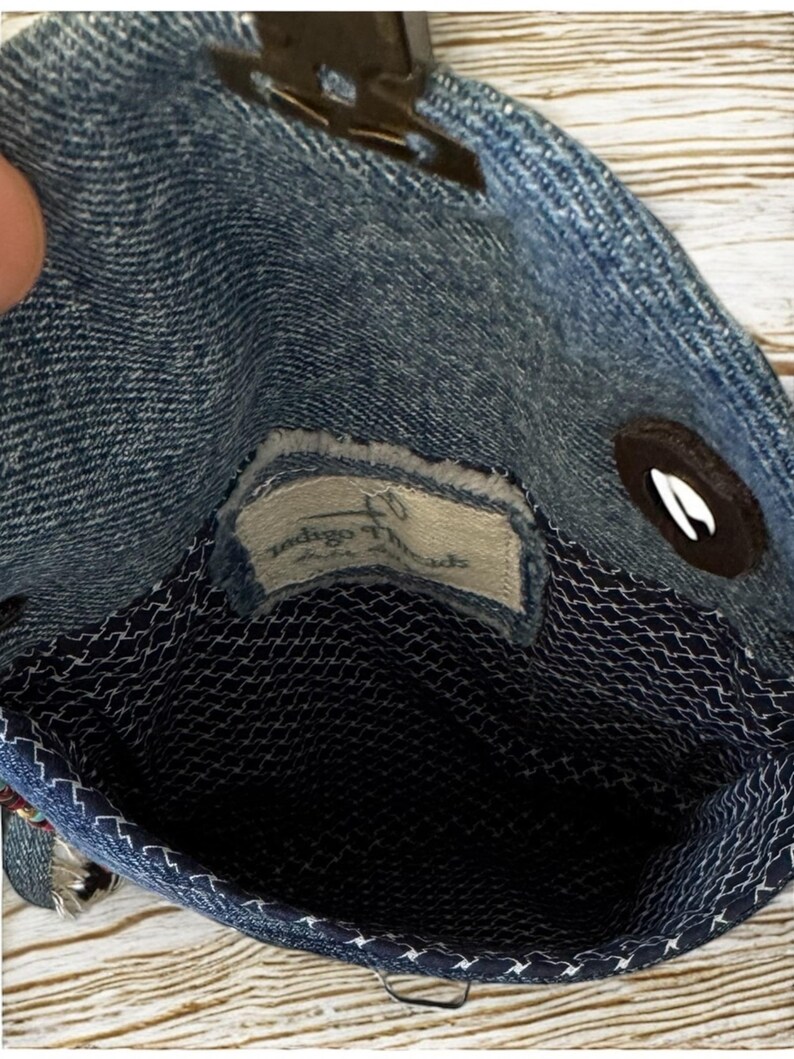 Op de afbeelding: Een denim handtas met een donkerblauwe binnenkant en een wit gestikt patroon. De tas heeft een magnetische sluiting en een klein label met de tekst "Indigo Threads". De tas is gemaakt van blauwe denim stof.