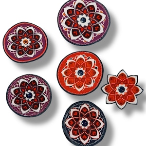 May include: A collection of decorative floral-patterned coasters in various colors, including red, purple, and orange. Each coaster features a central flower design with a button in the middle. The coasters are round and one is flower-shaped.