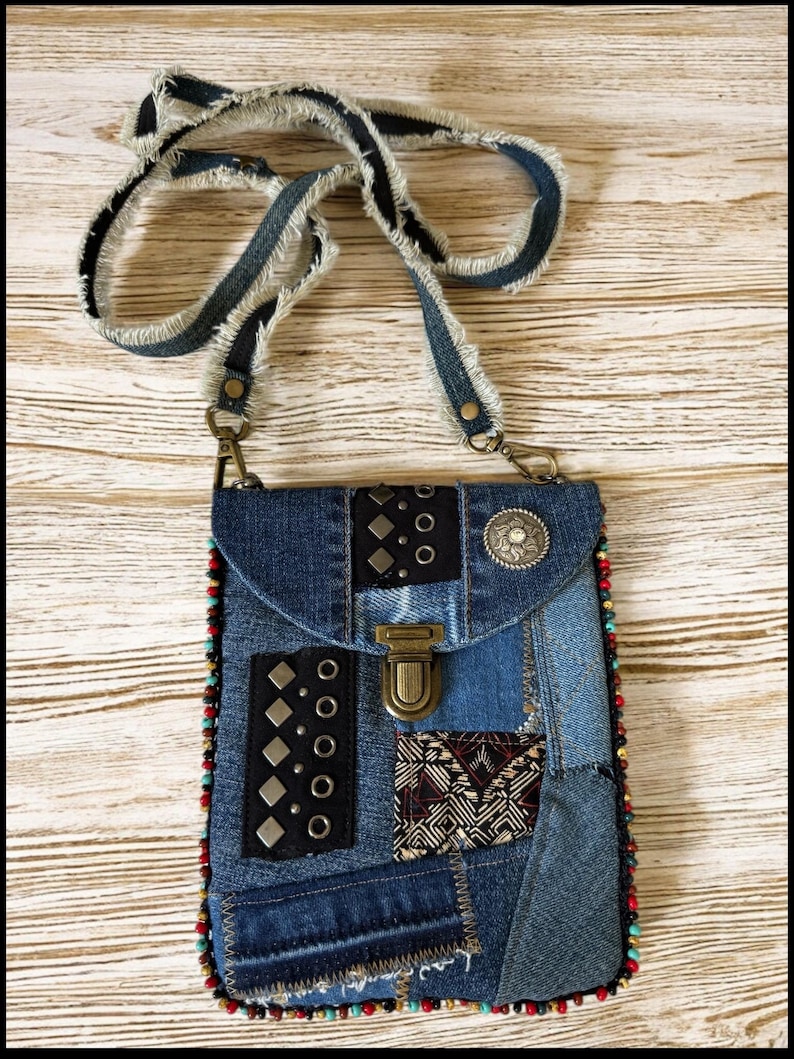 Op de afbeelding: Een denim patchwork crossbody tas met een gerafelde band. De tas heeft verschillende denim patches, metalen studs en een decoratieve sluiting. De randen zijn afgewerkt met kleurrijke kralen en een zilverkleurige versiering op de flap.