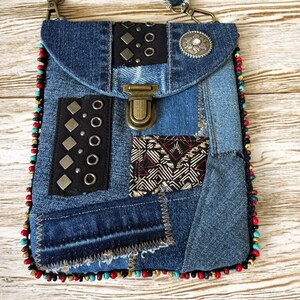Op de afbeelding: Een denim crossbody tas met een patchwork design. De tas heeft een klepsluiting, metalen accenten en een kralenrand. De riem is gemaakt van denim met rafelige randen. De tas is ongeveer 20 cm hoog en 15 cm breed.
