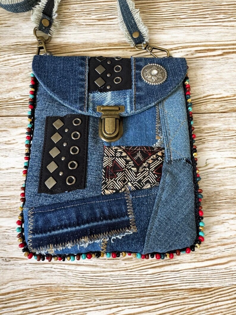 Op de afbeelding: Een denim crossbody tas met een patchwork design. De tas heeft een klepsluiting, metalen accenten en een kralenrand. De riem is gemaakt van denim met rafelige randen. De tas is ongeveer 20 cm hoog en 15 cm breed.