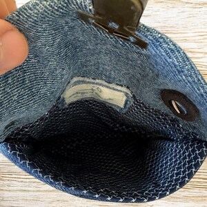 Op de afbeelding: Een kleine denim pouch met een metalen sluiting. De binnenkant is een donkerblauwe stof met een wit geometrisch patroon. De pouch is open en onthult de binnenkant. De denim is lichtblauw.