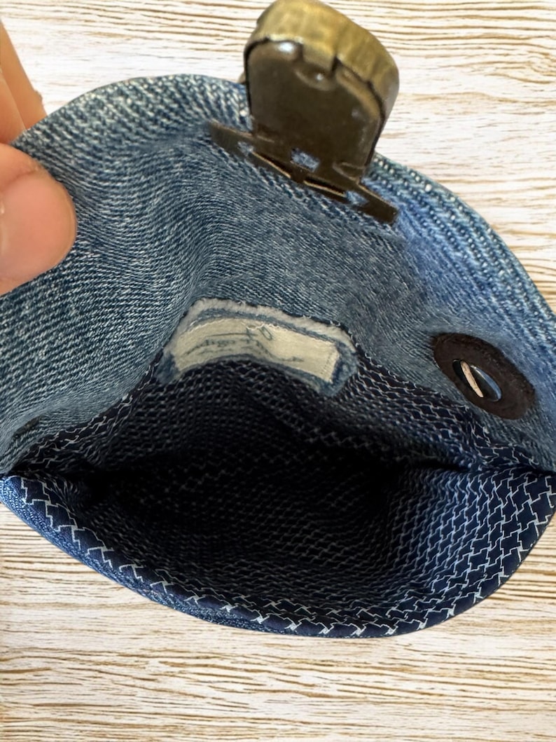 Op de afbeelding: Een kleine denim pouch met een metalen sluiting. De binnenkant is een donkerblauwe stof met een wit geometrisch patroon. De pouch is open en onthult de binnenkant. De denim is lichtblauw.