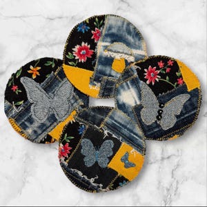May include: Four round fabric coasters with a patchwork design featuring denim, floral fabric, and butterflies. The coasters are yellow, black, and blue.