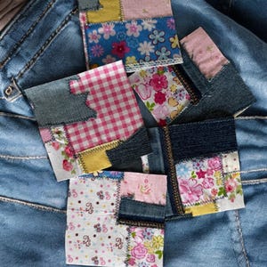 May include: Five small patchwork squares made of denim and floral fabric. The squares are arranged in a stack on a pair of blue jeans.