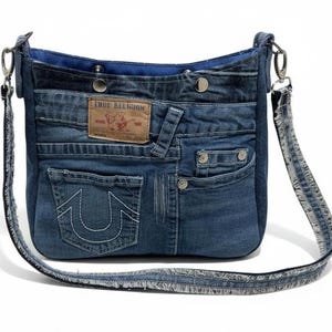 May include: A denim crossbody bag with a blue interior and a long, adjustable strap. The bag is constructed from repurposed jeans, featuring pockets, rivets, and a leather patch with the text "TRUE RELIGION". The bag has a casual, everyday style.
