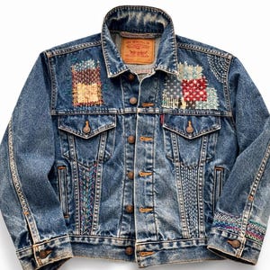 May include: A blue denim jacket with patches and decorative stitching. The jacket features a classic collar, button-down front, and two chest pockets. Patches include geometric designs in red, white, and blue. The sleeves have decorative stitching.