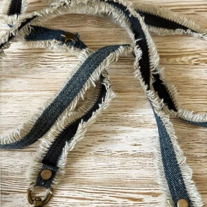 Op de afbeelding: Een denim tasriem met een gerafeld randontwerp. De riem is donkerblauw denim met een zwarte middenstreep en tan franjes. De riem heeft messingkleurige metalen hardware, inclusief sluitingen en klinknagels.