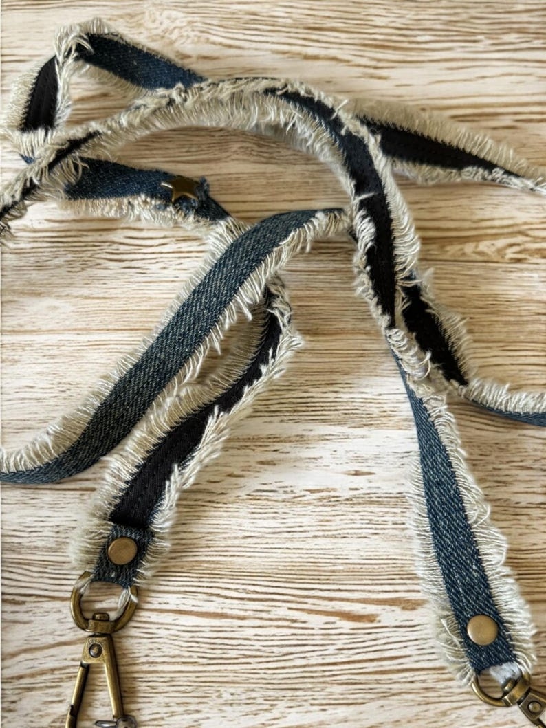 Op de afbeelding: Een denim tasriem met een gerafeld randontwerp. De riem is donkerblauw denim met een zwarte middenstreep en tan franjes. De riem heeft messingkleurige metalen hardware, inclusief sluitingen en klinknagels.