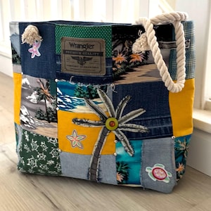 May include: A patchwork tote bag with a tropical theme. The bag features denim, yellow, and patterned fabric squares. It has a rope handle and decorative patches of a palm tree, starfish, and a turtle. A Wrangler label is visible.
