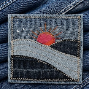 May include: A square denim patch with a stitched landscape scene. The design features a rising sun in shades of pink and orange, set against a blue sky. Below, a mountain range and a road are depicted in various shades of blue denim.