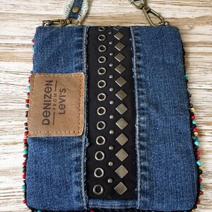 Op de afbeelding: Een denim crossbody tas met een zwart middenpaneel versierd met zilveren studs en grommets. De tas heeft een bruine leren patch met het "Denizen from Levi's" logo en is afgewerkt met kleurrijke kralen. De riem is gemaakt van denim.