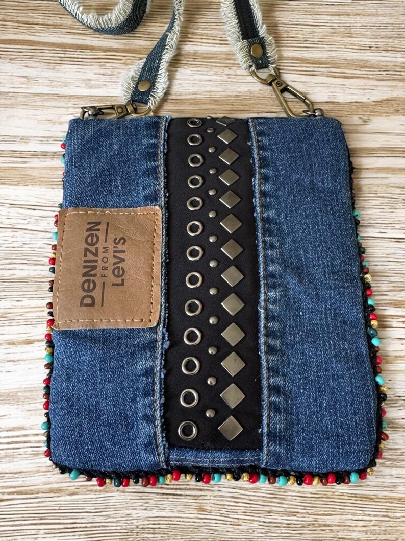 Op de afbeelding: Een denim crossbody tas met een zwart middenpaneel versierd met zilveren studs en grommets. De tas heeft een bruine leren patch met het "Denizen from Levi's" logo en is afgewerkt met kleurrijke kralen. De riem is gemaakt van denim.